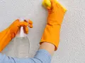 Mani con guanti arancioni puliscono un muro esterno con cappotto, usando uno spray e una spugna gialla.