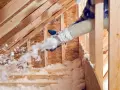 Lavoratore con guanti effettua insufflaggio sottotetto con materiale isolante bianco sparato da un tubo.