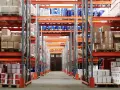 Magazzino con scaffalature arancioni piene di merci, tra cui barili blu e contenitori bianchi. Un corridoio centrale conduce a una porta doppia. Categoria catastale c/2.