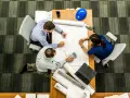 Team di architetti discute progetti su un tavolo, con rotoli di disegni e un casco blu. L'ufficio, con moquette a righe, è ideale per una **categoria catastale A10**.