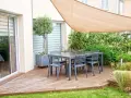 Un tavolo con sedie su un pavimento da giardino su terra, con un ulivo in vaso e una vela parasole.
