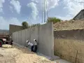 Operai installano un grande muro di contenimento in cemento armato, sollevato da una gru. Il cantiere è in pendenza.