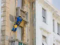 Operaio in corda ripara la facciata di un palazzo, lavorando sui dettagli dei balconi aggettanti.