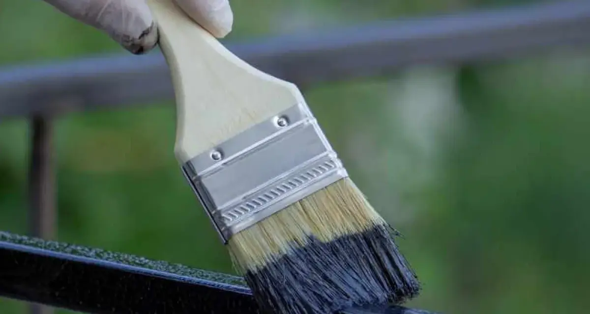 Mano con guanto bianco applica vernice scura su ringhiera metallica con pennello, un modo rapido per verniciare ringhiera senza carteggiare.