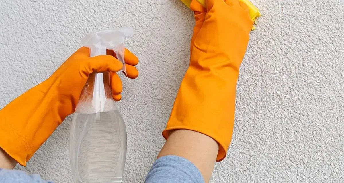 Mani con guanti arancioni puliscono un muro esterno con cappotto, usando uno spray e una spugna gialla.