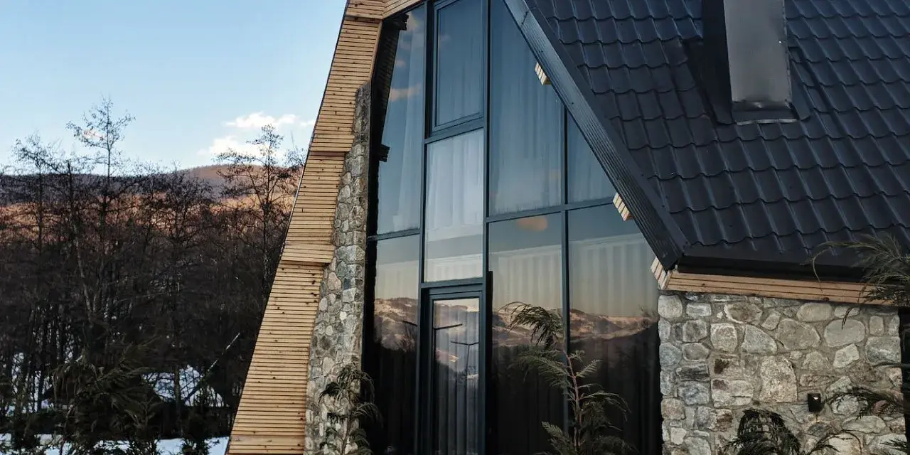 Moderna ristrutturazione casa di montagna: tetto scuro, ampie vetrate con vista sui monti, pietra e legno.