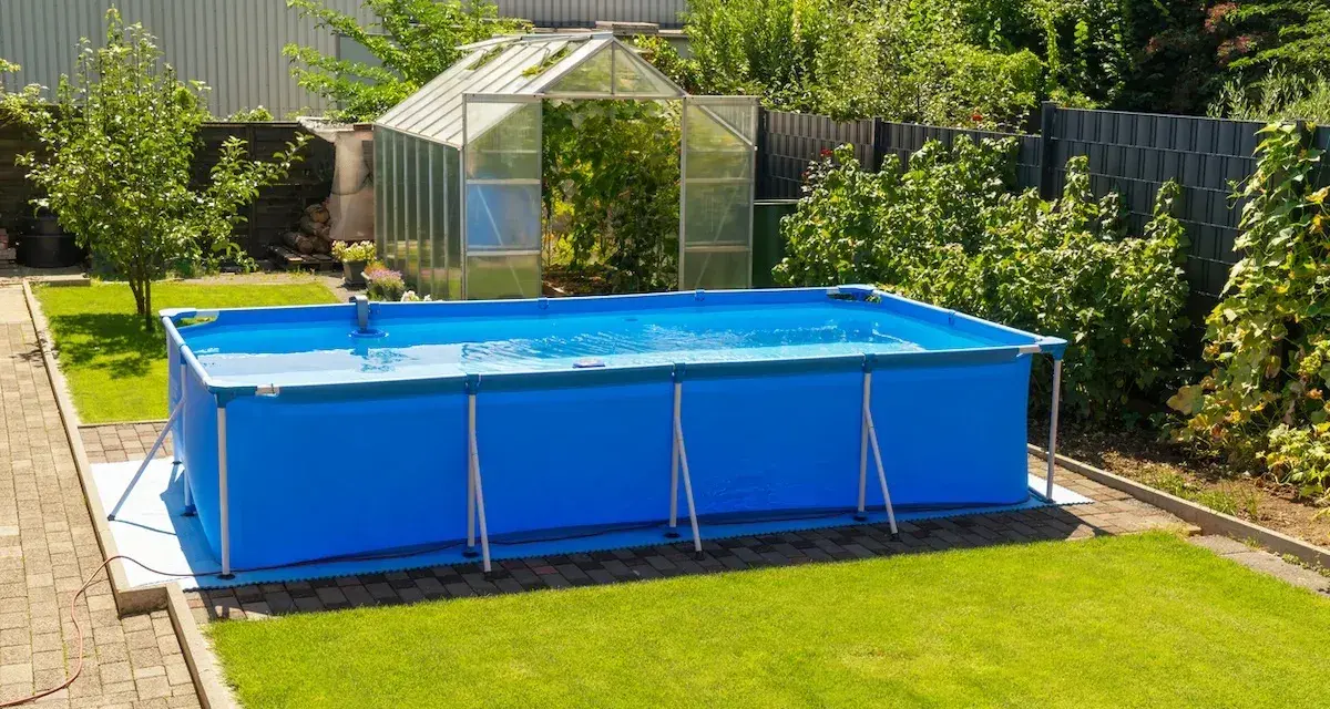 Ampia piscina blu montata in giardino, con serra sullo sfondo. Sembra una piscina senza permessi, pronta per l'estate.