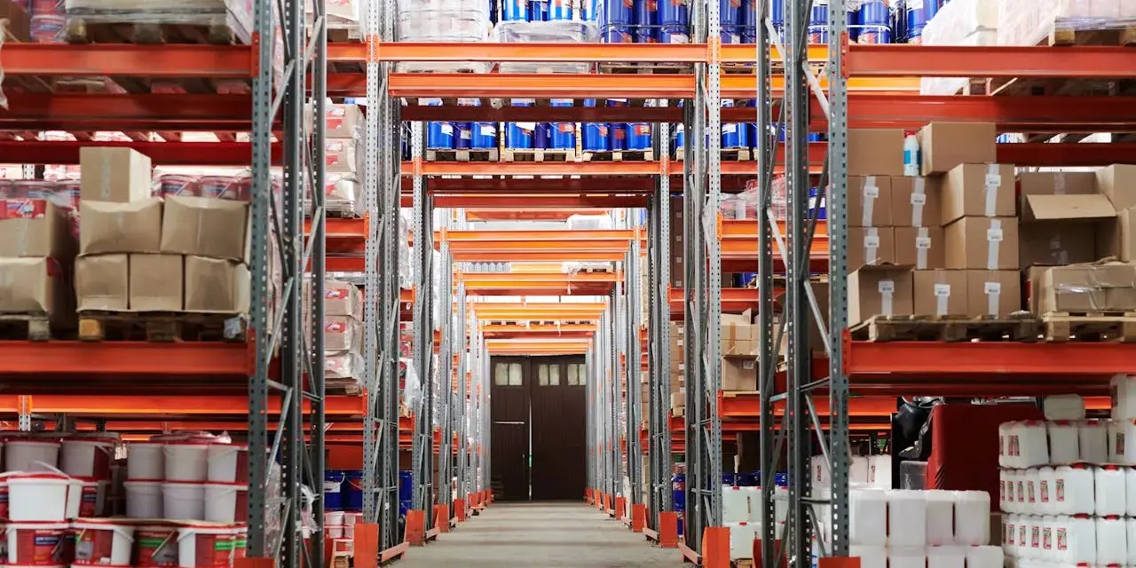 Magazzino con scaffalature arancioni piene di merci, tra cui barili blu e contenitori bianchi. Un corridoio centrale conduce a una porta doppia. Categoria catastale c/2.