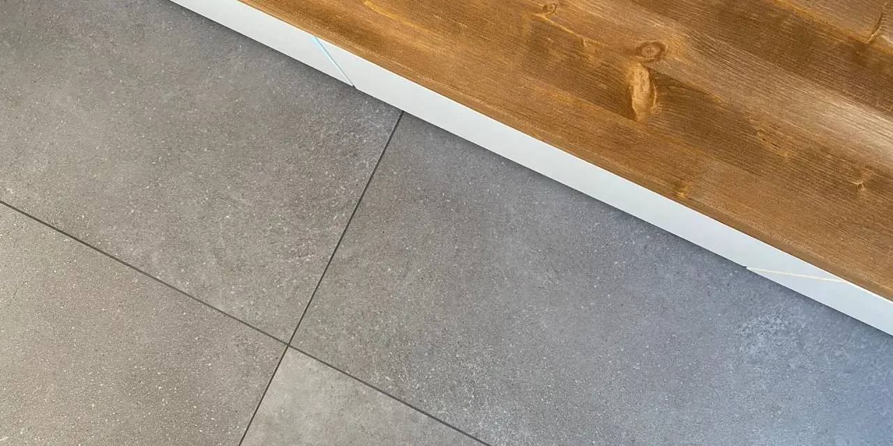 Dettaglio di piastrelle grigie posate con precisione, pronte per attaccare piastrelle al muro con silicone. Sopra, una trave di legno grezzo.
