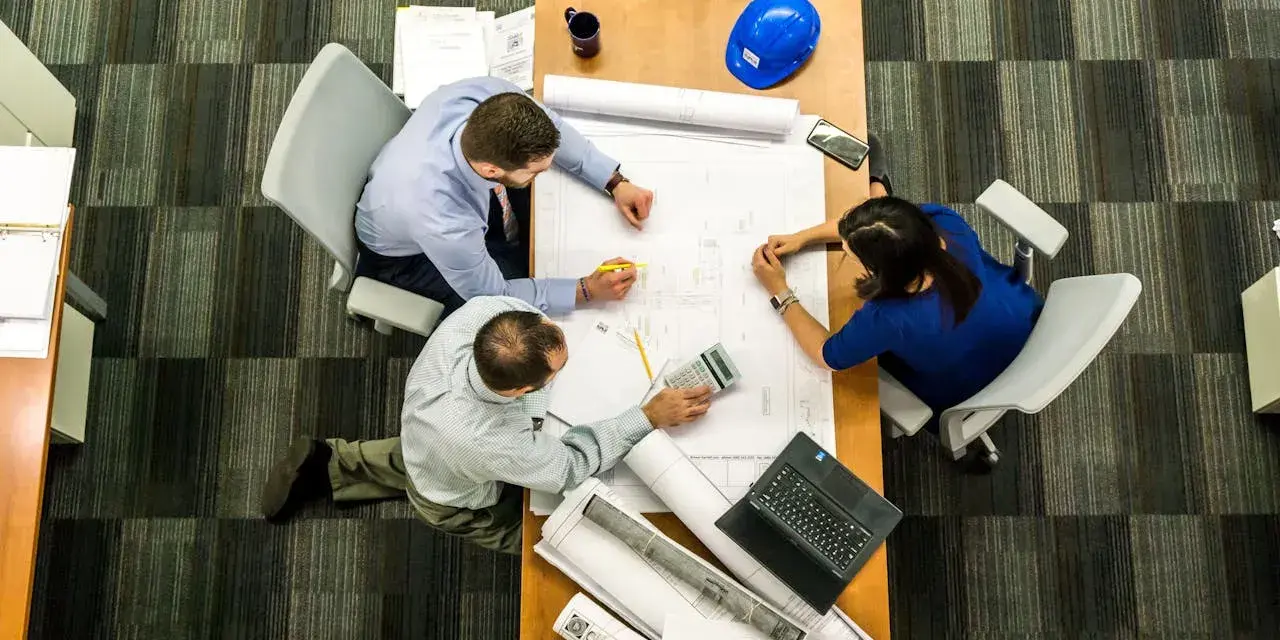 Team di architetti discute progetti su un tavolo, con rotoli di disegni e un casco blu. L'ufficio, con moquette a righe, è ideale per una **categoria catastale A10**.