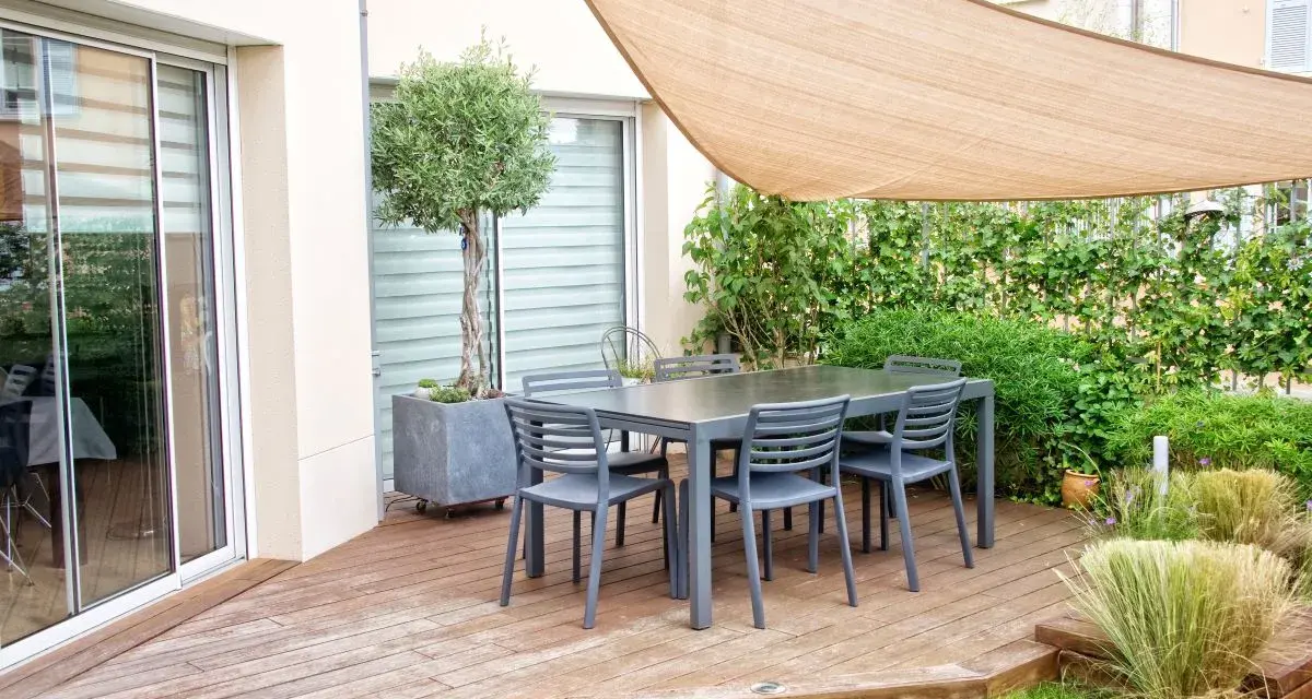 Un tavolo con sedie su un pavimento da giardino su terra, con un ulivo in vaso e una vela parasole.