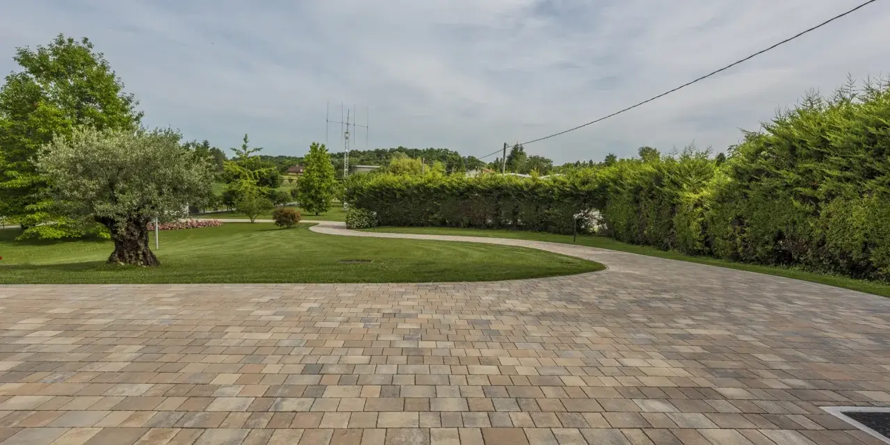 Pavimentazione giardino a secco con blocchi color sabbia, che conducono a un prato verde curato con alberi e siepi.