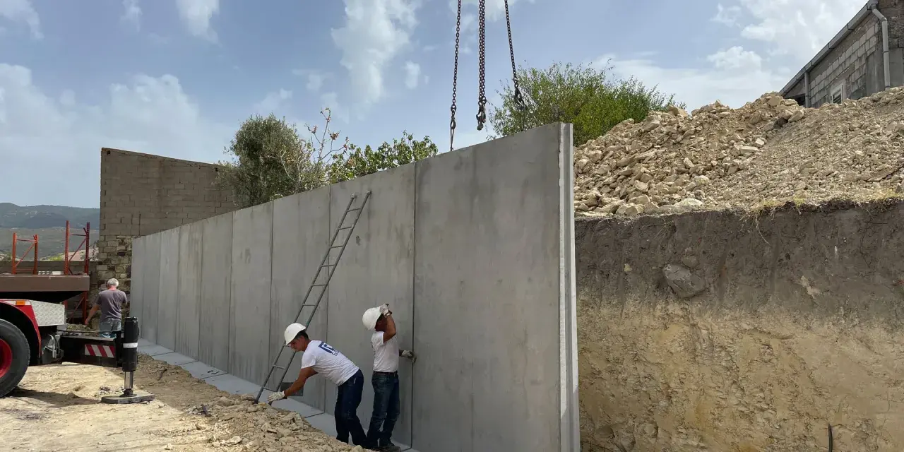 Operai installano un grande muro di contenimento in cemento armato, sollevato da una gru. Il cantiere è in pendenza.