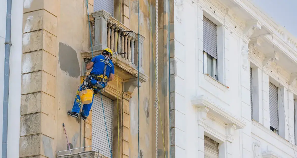 Operaio in corda ripara la facciata di un palazzo, lavorando sui dettagli dei balconi aggettanti.