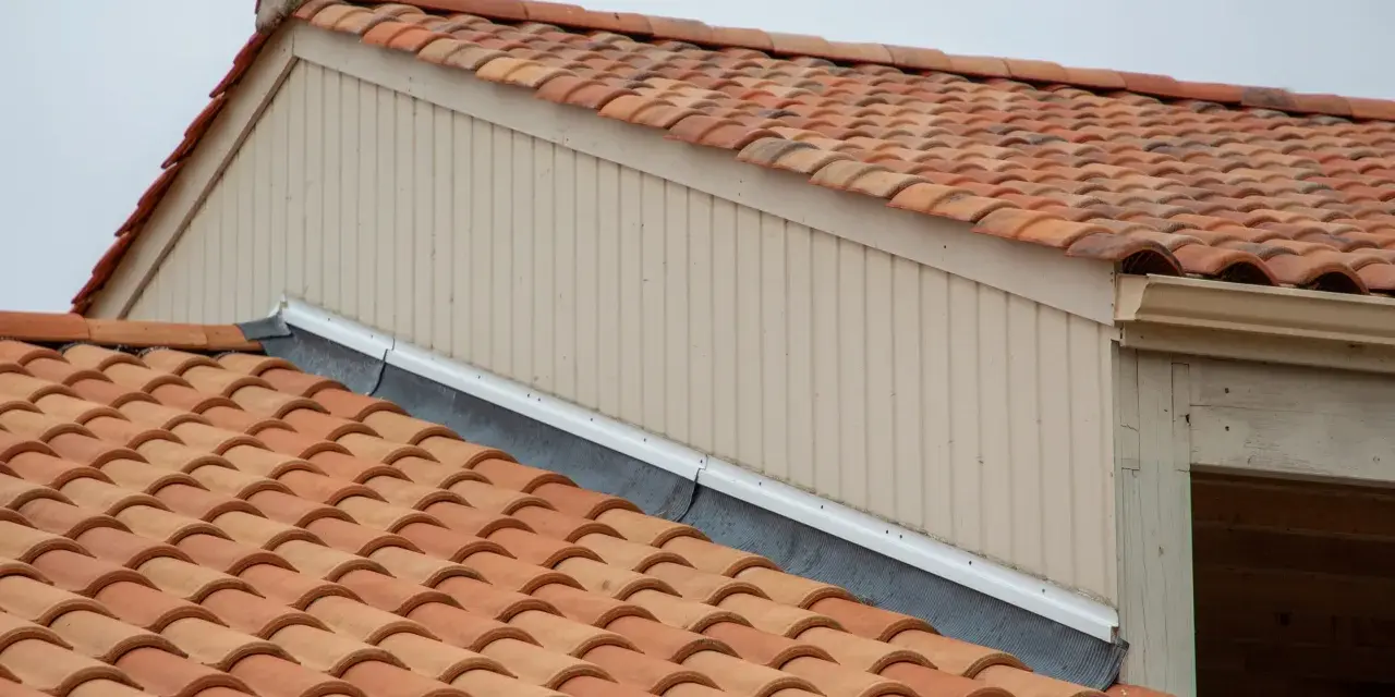 Tegole rosse su un tetto, con pannelli verticali beige e una grondaia. Le scossaline proteggono il bordo del tetto.