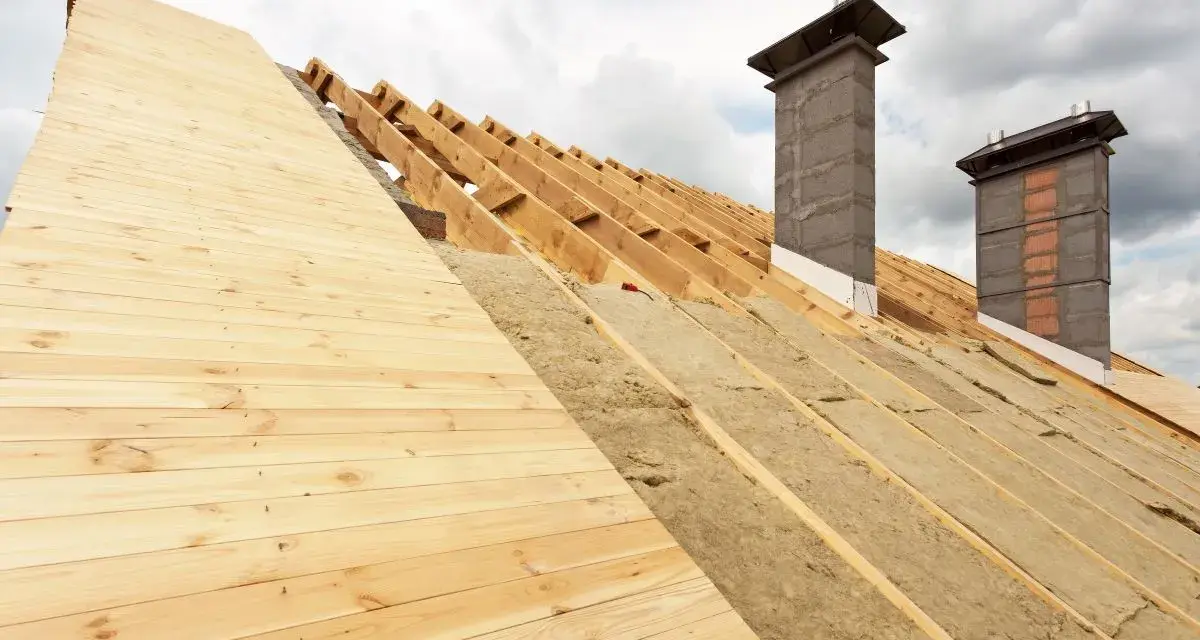 Costruzione tetto con isolamento termico: legno, lana di roccia e camini in muratura sotto un cielo nuvoloso.