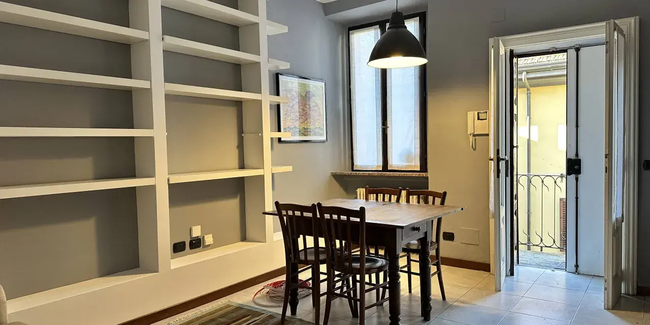 Un soggiorno con libreria a giorno, tavolo in legno e sedie. Dalla porta si intravede un cortile, tipico delle case di ringhiera.