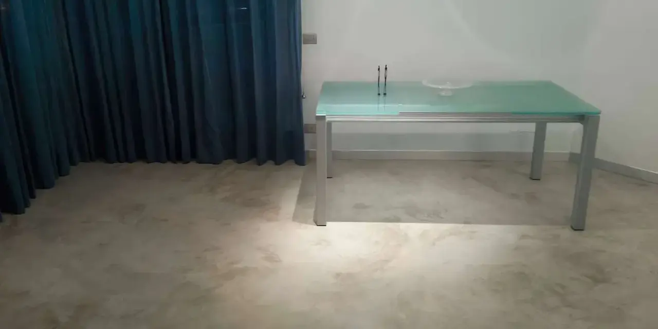 Un tavolo moderno con piano in vetro poggia su un **pavimento in cemento grezzo** dall'aspetto materico, con tende blu sullo sfondo.