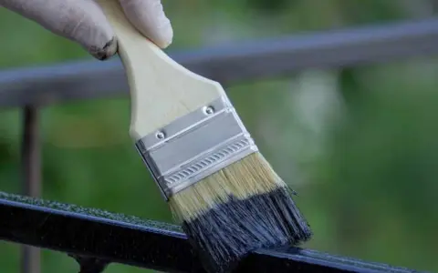 Mano con guanto bianco applica vernice scura su ringhiera metallica con pennello, un modo rapido per verniciare ringhiera senza carteggiare.