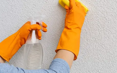 Mani con guanti arancioni puliscono un muro esterno con cappotto, usando uno spray e una spugna gialla.