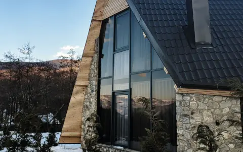 Moderna ristrutturazione casa di montagna: tetto scuro, ampie vetrate con vista sui monti, pietra e legno.