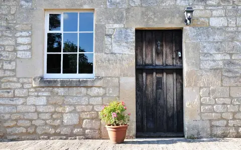 Facciate in pietra e mattoni grezzi, con una porta in legno scuro e una finestra bianca. Un vaso di fiori rosa aggiunge un tocco di colore.