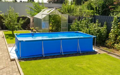 Ampia piscina blu montata in giardino, con serra sullo sfondo. Sembra una piscina senza permessi, pronta per l'estate.