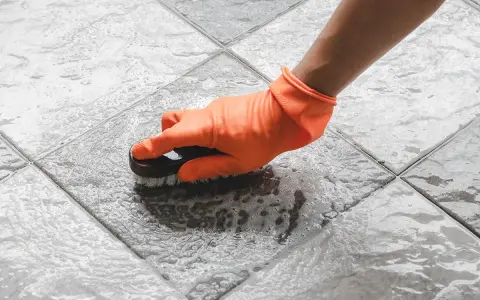 Mano con guanto arancione che spazzola un pavimento piastrellato bagnato, mostrando come togliere ruggine dal pavimento con schiuma.