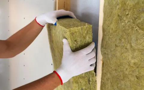 Mani guantate posizionano un pannello isolante, creando un cappotto interno o esterno per una parete.