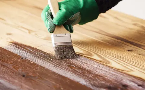 Mano guantata applica vernice protettiva con pennello per impermeabilizzare il legno, creando una finitura lucida.