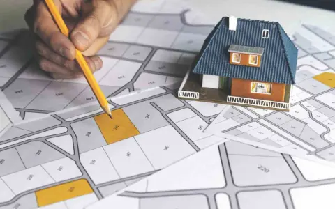 Mano con matita su mappa catastale, con modellino di casa. Pianificazione immobiliare, categoria catastale D10.