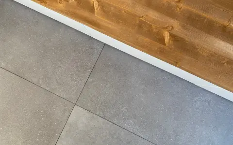 Dettaglio di piastrelle grigie posate con precisione, pronte per attaccare piastrelle al muro con silicone. Sopra, una trave di legno grezzo.