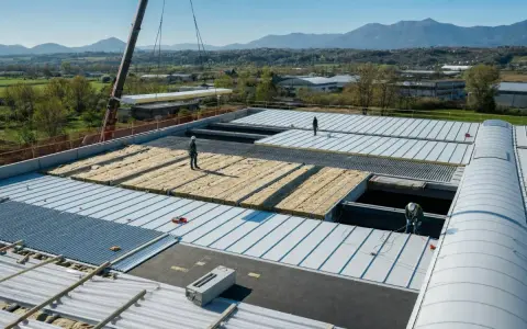 Operai installano pannelli isolanti su un tetto, riducendo il rumore. Un cantiere in corso con montagne sullo sfondo.
