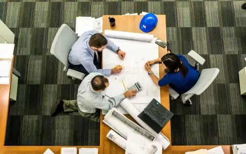 Team di architetti discute progetti su un tavolo, con rotoli di disegni e un casco blu. L'ufficio, con moquette a righe, è ideale per una **categoria catastale A10**.