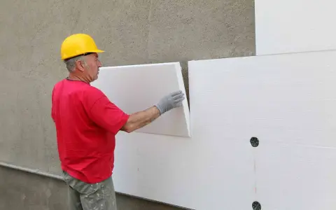 Operaio con casco giallo applica pannelli isolanti bianchi. Attenzione: il polistirolo è infiammabile.