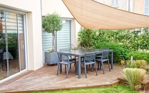 Un tavolo con sedie su un pavimento da giardino su terra, con un ulivo in vaso e una vela parasole.
