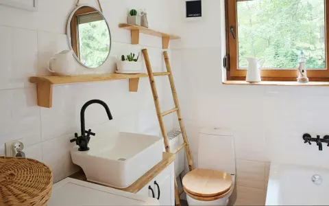 Bagno piccolo con piastrelle bianche lucide, lavabo squadrato, specchio rotondo e scala in bambù. Idee piastrelle bagno piccolo: la luminosità è la chiave.