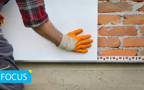 Installazione battiscopa per cappotto esterno: un operaio con guanti arancioni posiziona un pannello isolante bianco su una parete di mattoni, con un profilo metallico alla base.