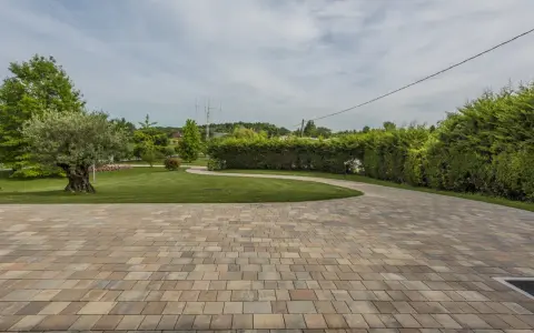 Pavimentazione giardino a secco con blocchi color sabbia, che conducono a un prato verde curato con alberi e siepi.