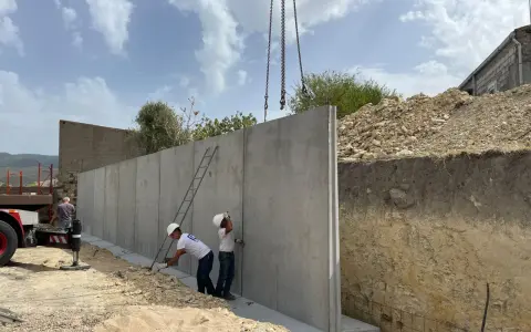 Operai installano un grande muro di contenimento in cemento armato, sollevato da una gru. Il cantiere è in pendenza.