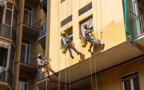 Operai in corda applicano diversi tipi di intonaco per esterni su un edificio giallo.