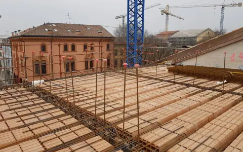 Costruzione di un solaio: mattoni forati e armature in ferro pronti per il getto di cemento. In lontananza, un edificio storico e gru edili.