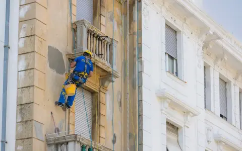 Operaio in corda ripara la facciata di un palazzo, lavorando sui dettagli dei balconi aggettanti.