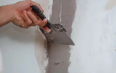 Mano che usa una spatola per stuccare un muro, creando una superficie liscia e uniforme per la pittura.