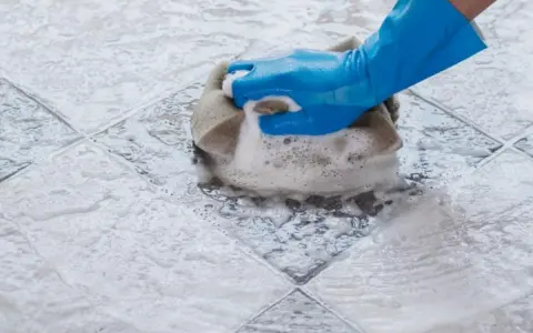 Mano con guanto blu che pulisce ceramica macchiata con spugna e schiuma.