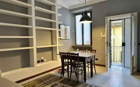 Un soggiorno con libreria a giorno, tavolo in legno e sedie. Dalla porta si intravede un cortile, tipico delle case di ringhiera.