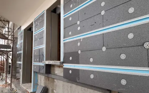 Pannelli grigi per cappotto termico ignifugo, fissati con tasselli bianchi e profili azzurri, in fase di installazione su un edificio.