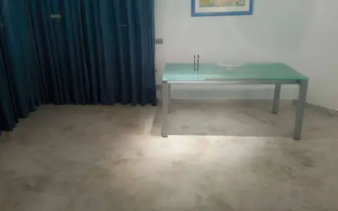 Un tavolo moderno con piano in vetro poggia su un **pavimento in cemento grezzo** dall'aspetto materico, con tende blu sullo sfondo.