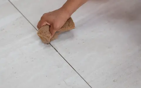 Mano che stuccare piastrelle con una spugna, rifinendo le fughe di un pavimento chiaro.