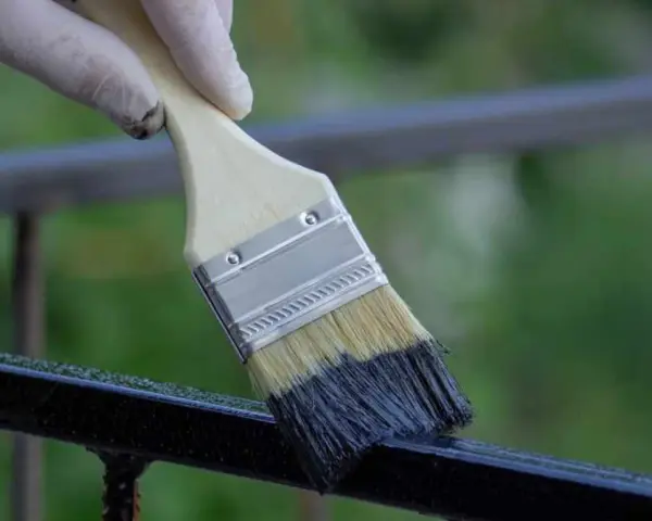 Mano con guanto bianco applica vernice scura su ringhiera metallica con pennello, un modo rapido per verniciare ringhiera senza carteggiare.